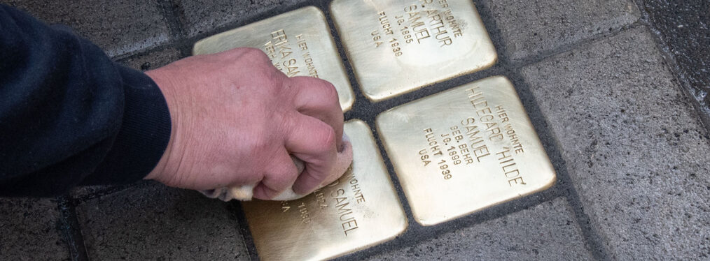 Eine Hand wischt mit einem Tuch über vier gerade verlegte Stolpersteine