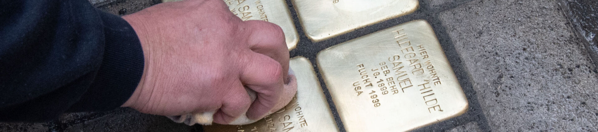 Eine Hand wischt mit einem Tuch über vier gerade verlegte Stolpersteine